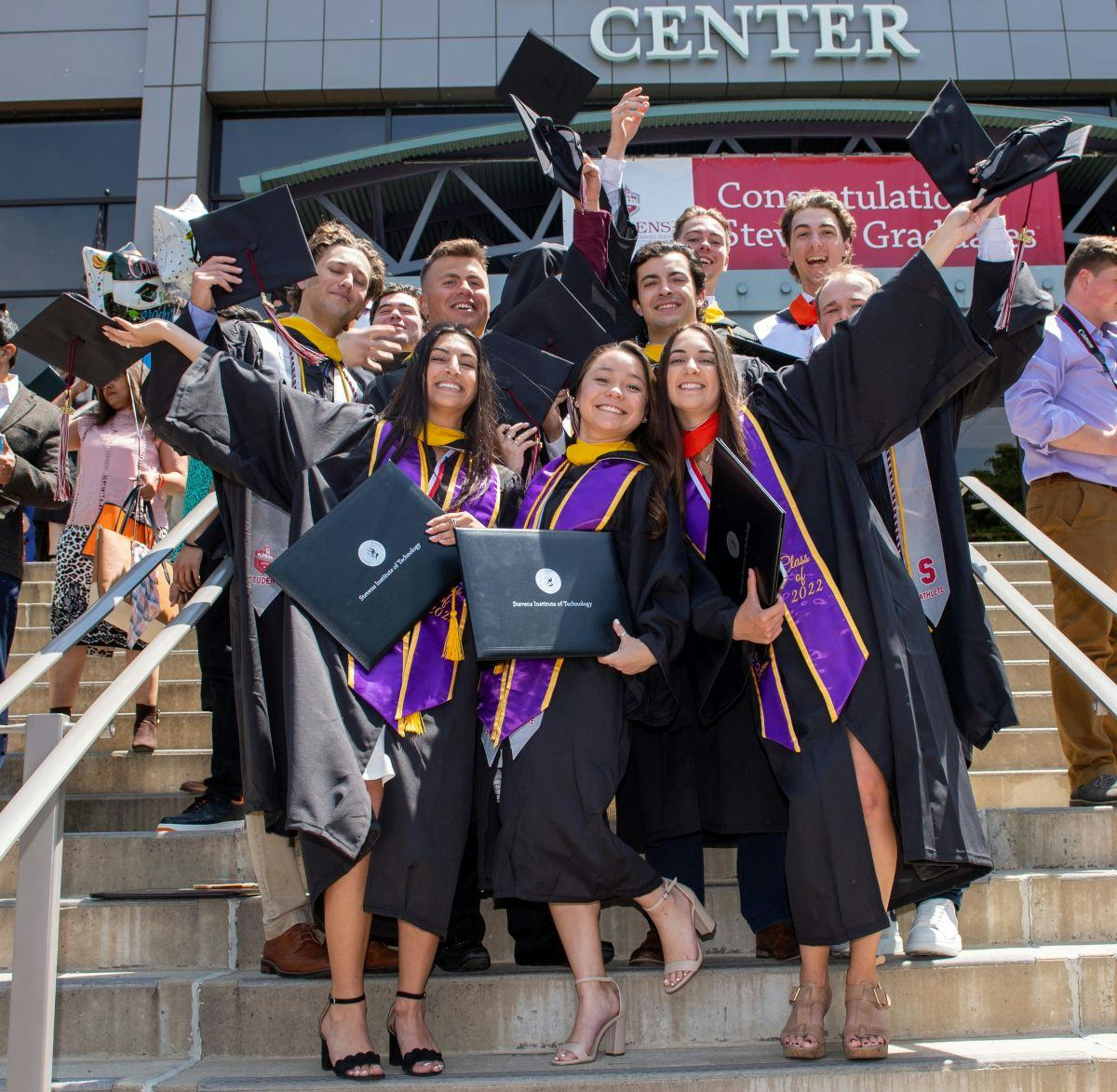 Students pose and smile in cap and gown, holding their diplomas, on the steps of Meadowlands Exposition Center