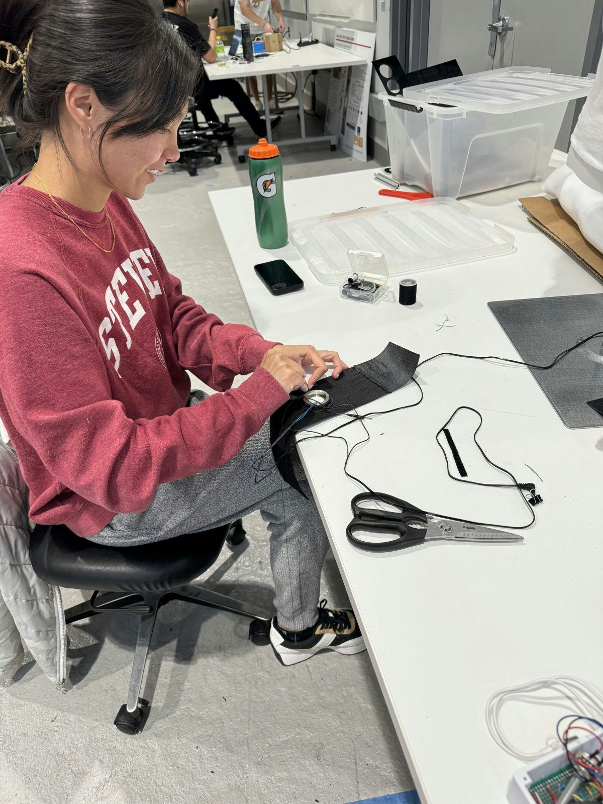 A female student in a red Stevens sweatshirt sits at a table in a lab with a pair of scissors next to her. In her hands she is working on assembling the Medflex device.