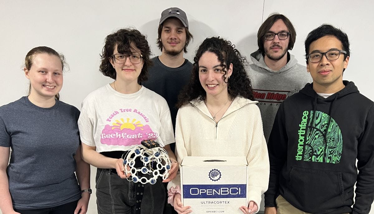 A group of six students stand together. One is holding an EEG cap that is placed over the subject's head, and another holds a box that says "OPEN BCI" on it.