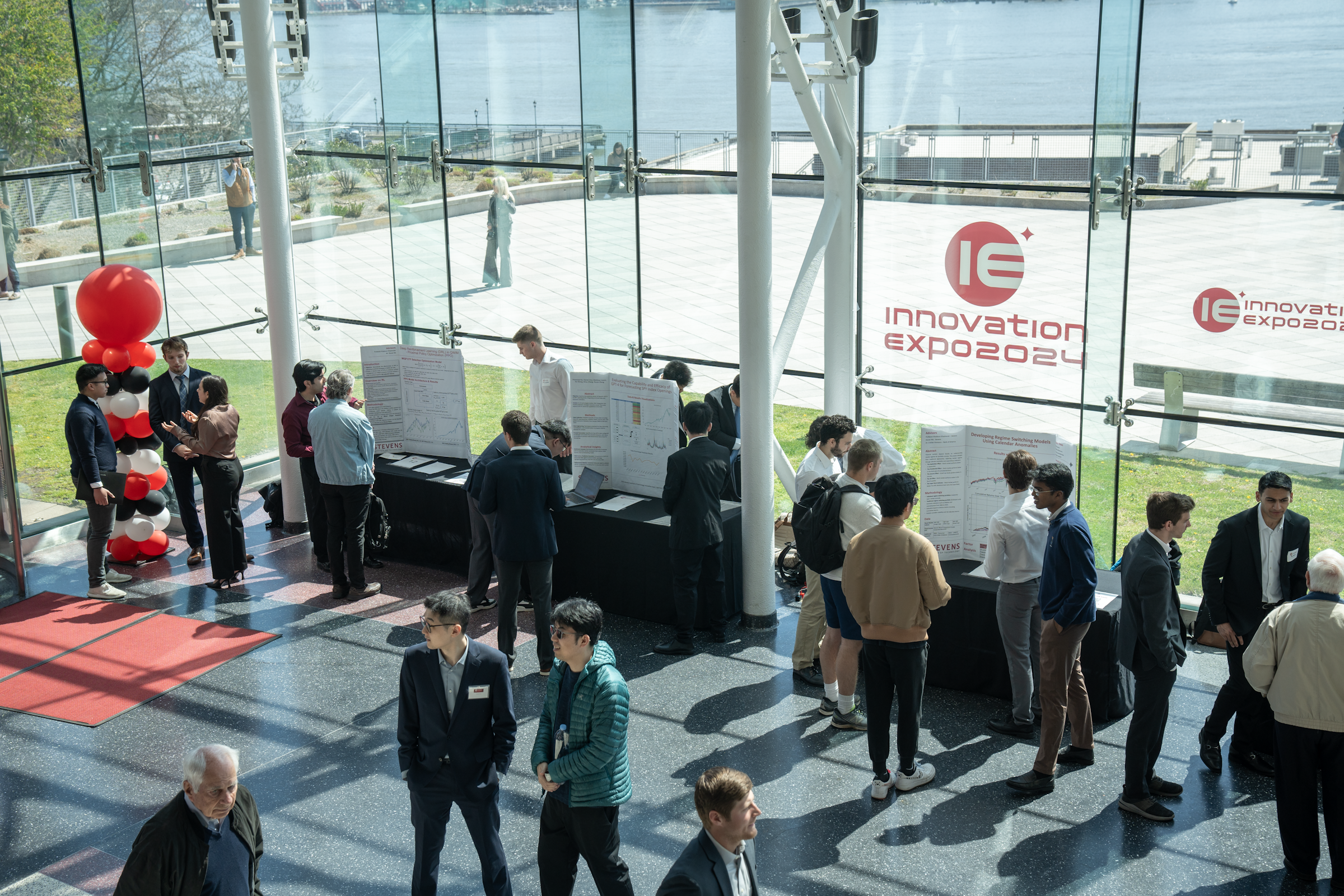 An overhead shot of students presenting their projects in the atrium of the Babbio Center in front of the glass wall with the Hudson River in the background. 