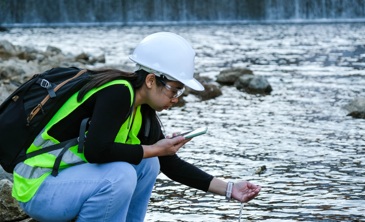 An environmental engineering student in a hard helmet and vest testing water samples with a phone.