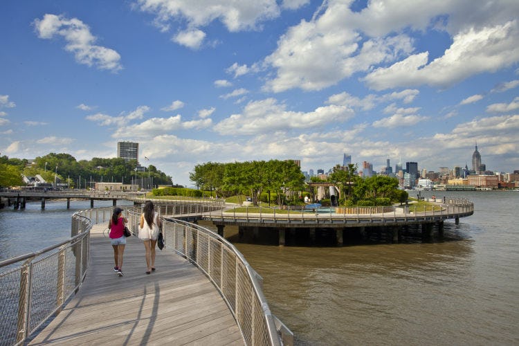 Pedestrians walking onto a pier park along Hoboken's waterside with New York in the background. 