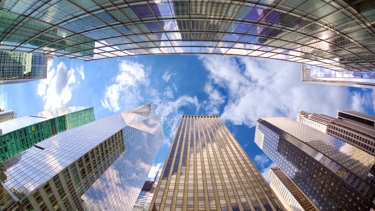 A view of midtown Manhattan skyscrapers from street level looking straight up into a blue sky with white clouds.