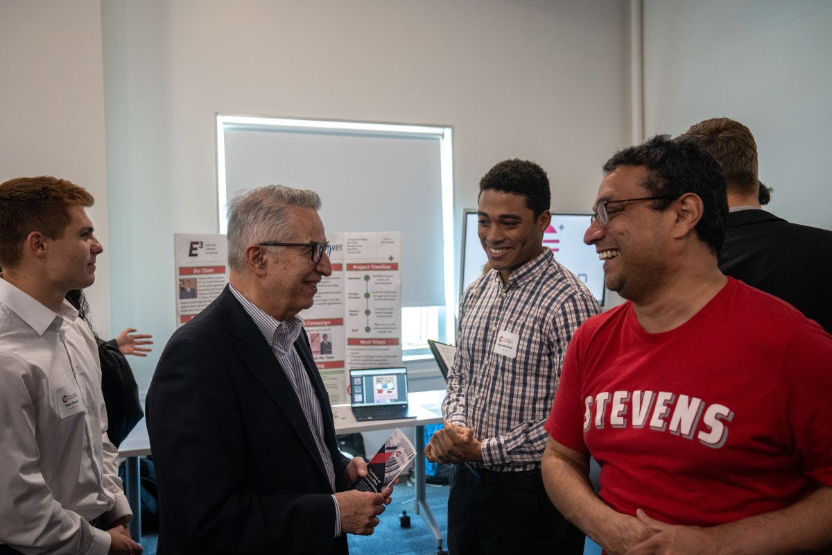 Gregory Prastacos stands on the left wearing a blue blazer talking to a student in the middle wearing a checkered shirt about his project.Professor Guarav Sabnis wearing a red Stevens t-shirt is to the right.
