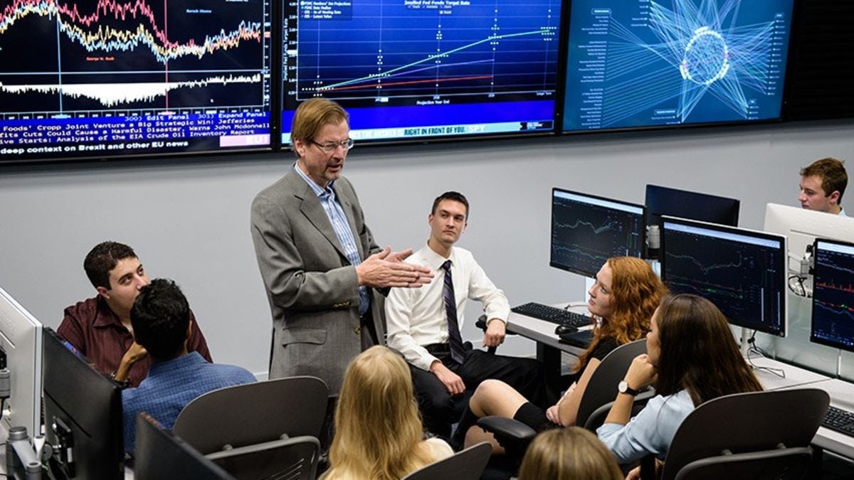 A professor teaches a class in the Hanlon Lab, with several screens behind him showing live financial market data.