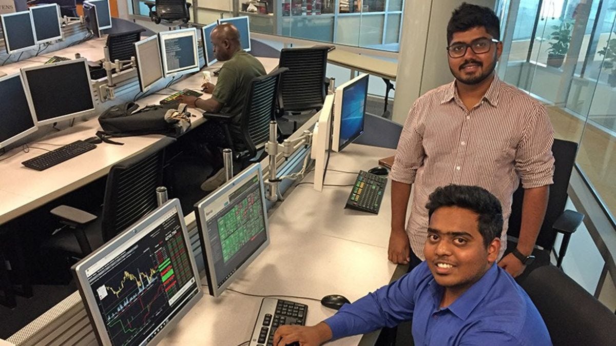 Vishnu Pillai, in a striped shirt, stands over Akshay Kumar Vikram, in a blue shirt, as he pulls up some data on a Bloomberg terminal at one of the high-tech workstations in a Stevens lab.