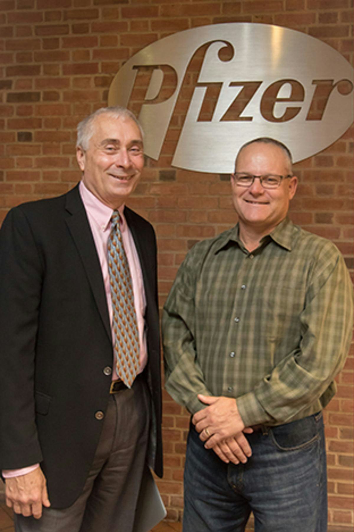 Two men in front of a brick wall with the Pfizer logo.