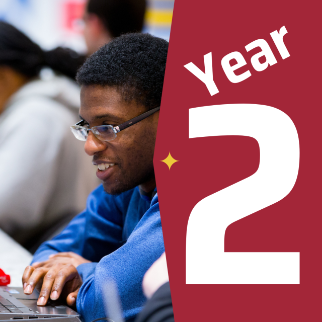 A male Black student wearing a blue shirt sitting at a table in front of a laptop next to a graphic that says year two.