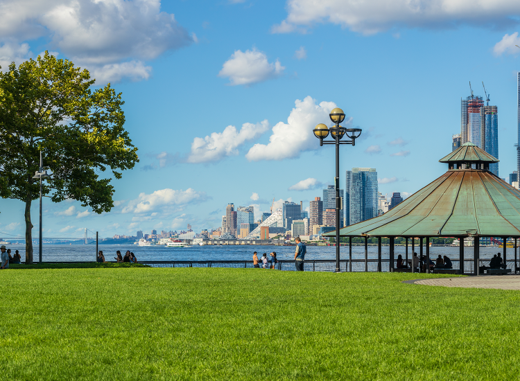 Hoboken Millennium Park features a tall green tree and a large covered space in the park, with part of the New York City skyline in the background.