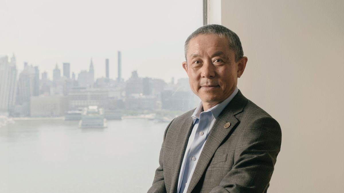 Provost Jianmin Qu poses for the camera in front of a window overlooking the New York skyline.