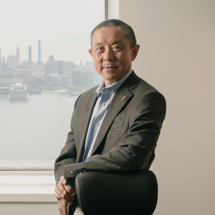 Provost Jianmin Qu poses for the camera in front of a window overlooking the New York skyline.