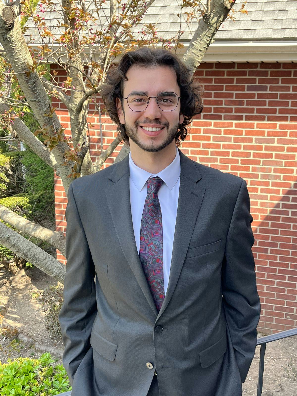 Mechanical engineering senior Drew Maggio ’23, wearing a suit and tie, smiles while standing outside in front of a tree.