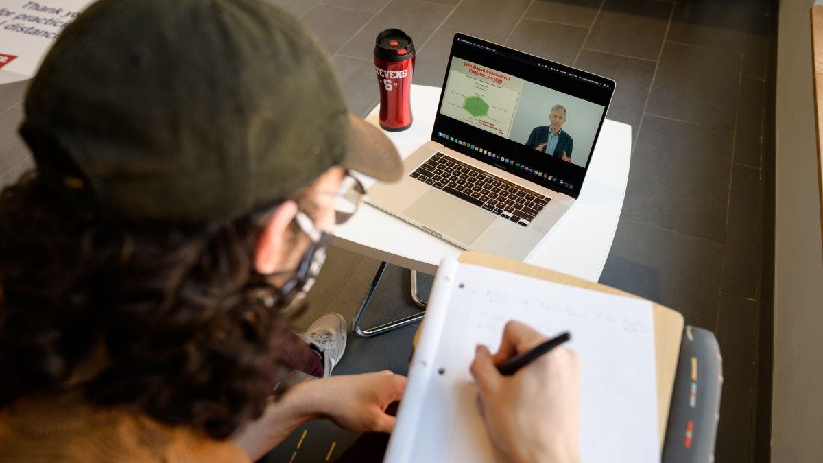 Student in ball cap writes notes while viewing Stevens lecture on laptop