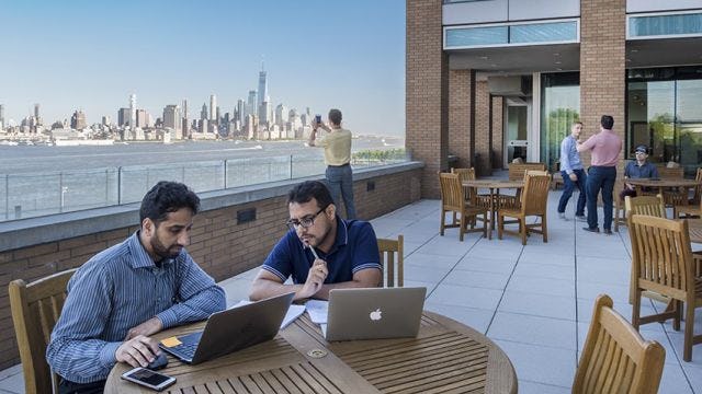 Groups of students working on a patio overlooking the lower Manhattan skyline. Two male students look at laptops in the foreground.