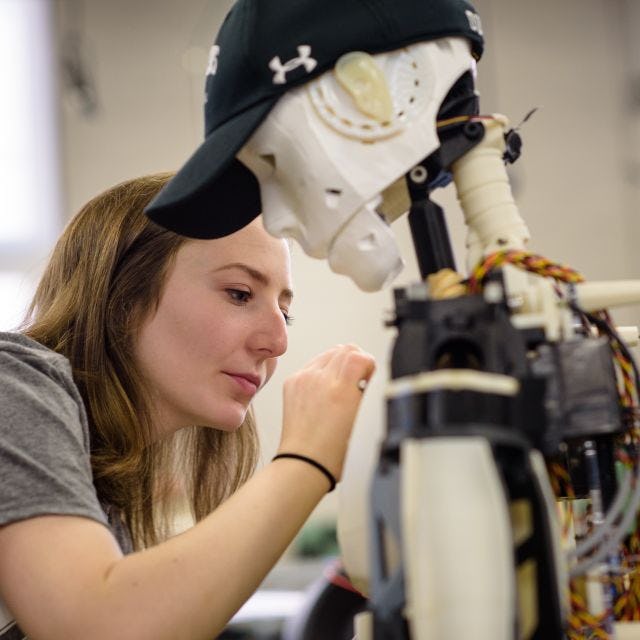 Student working on a white robot wearing a black Stevens hat with wires showing in its back