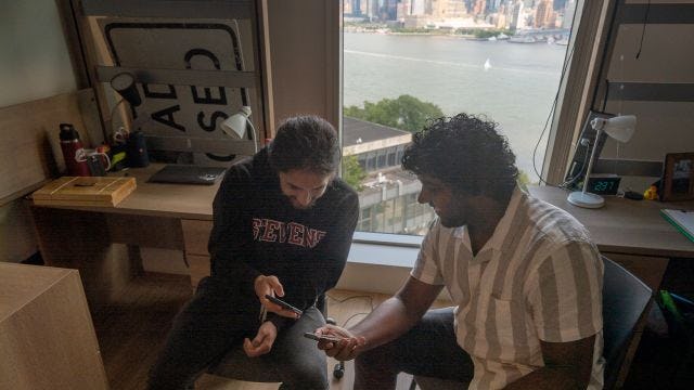 Students in dorm show each other their phones with New York city skyline through window behind them.