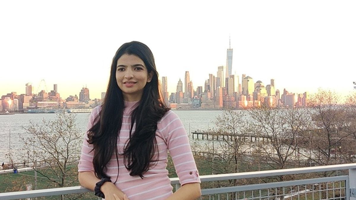 Aayushi Gandhi stands before the New York City skyline at sunset. The Hudson River is clearly visible in the background.