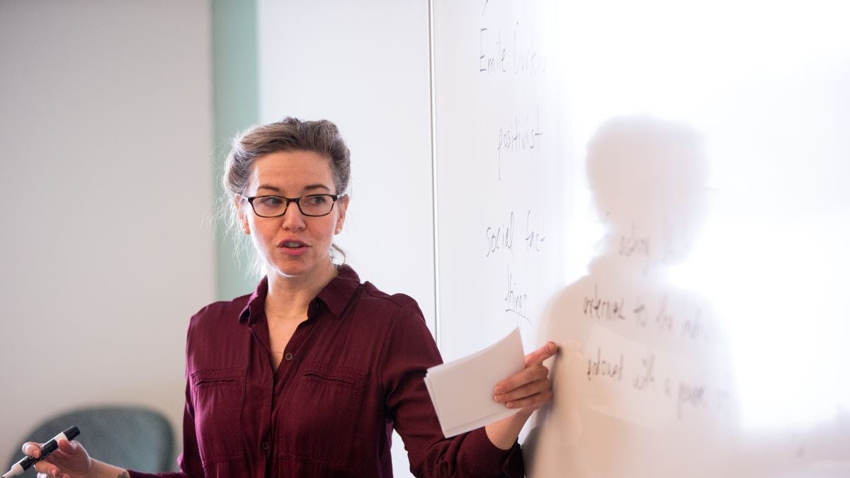 Professor in front of class gestures toward white board with sociology text written on it