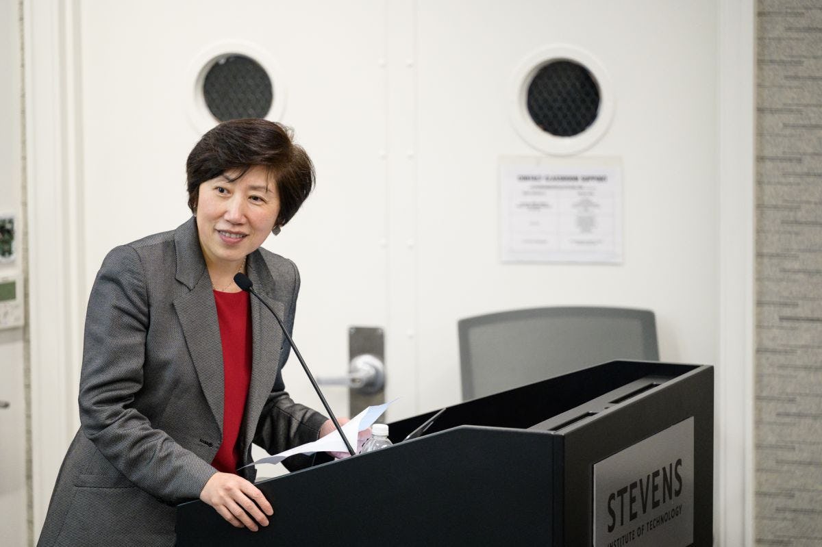 Jean Zu stands behind a podium over a microphone looking over her shoulder and speaking