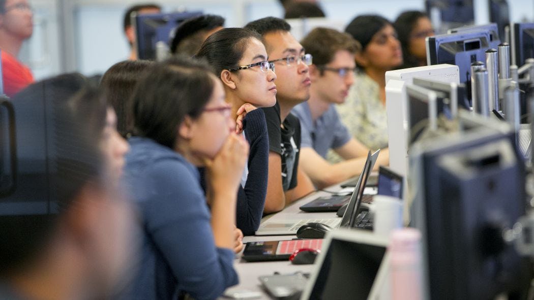 Students in front of computer monitors listen to instructor 