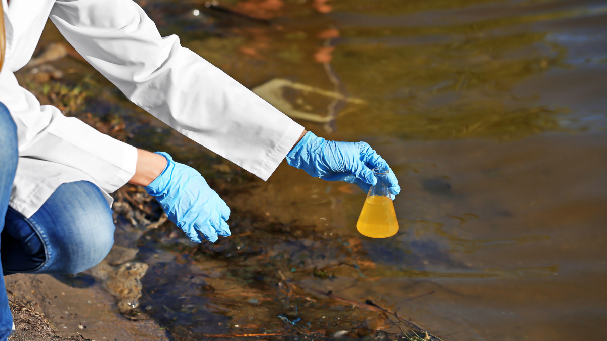 Unidentified person in lab coat and gloves holds beaker above body of water