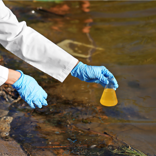 Unidentified person in lab coat and gloves holds beaker above body of water