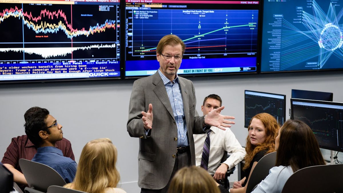 Professor speaks to group of students in front of monitors displaying finance data