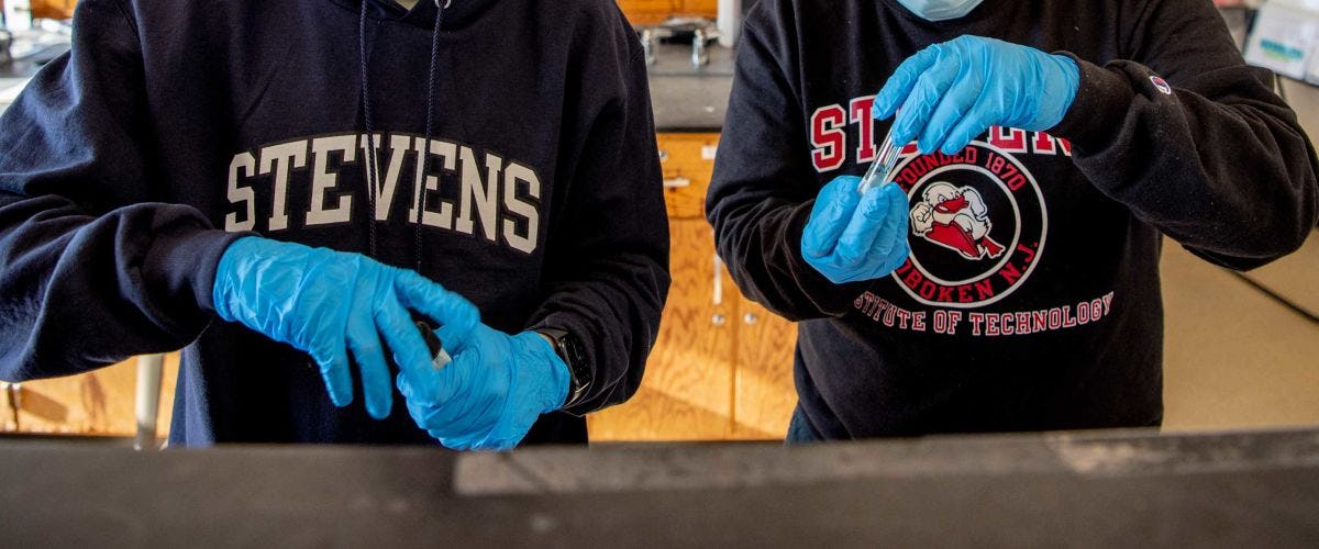 Two pairs of students gloved hands work with chemistry equipment while wearing Stevens apparel.