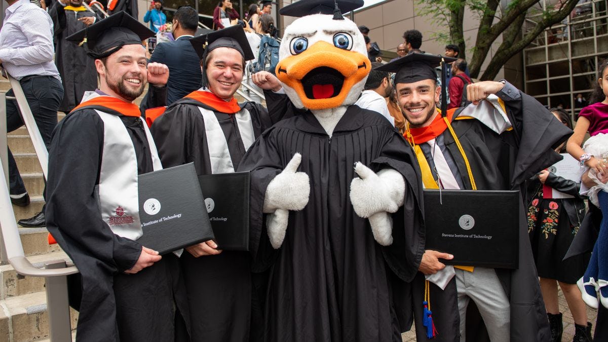 Attila the duck in cap and gown with thumbs up in group of recent graduates