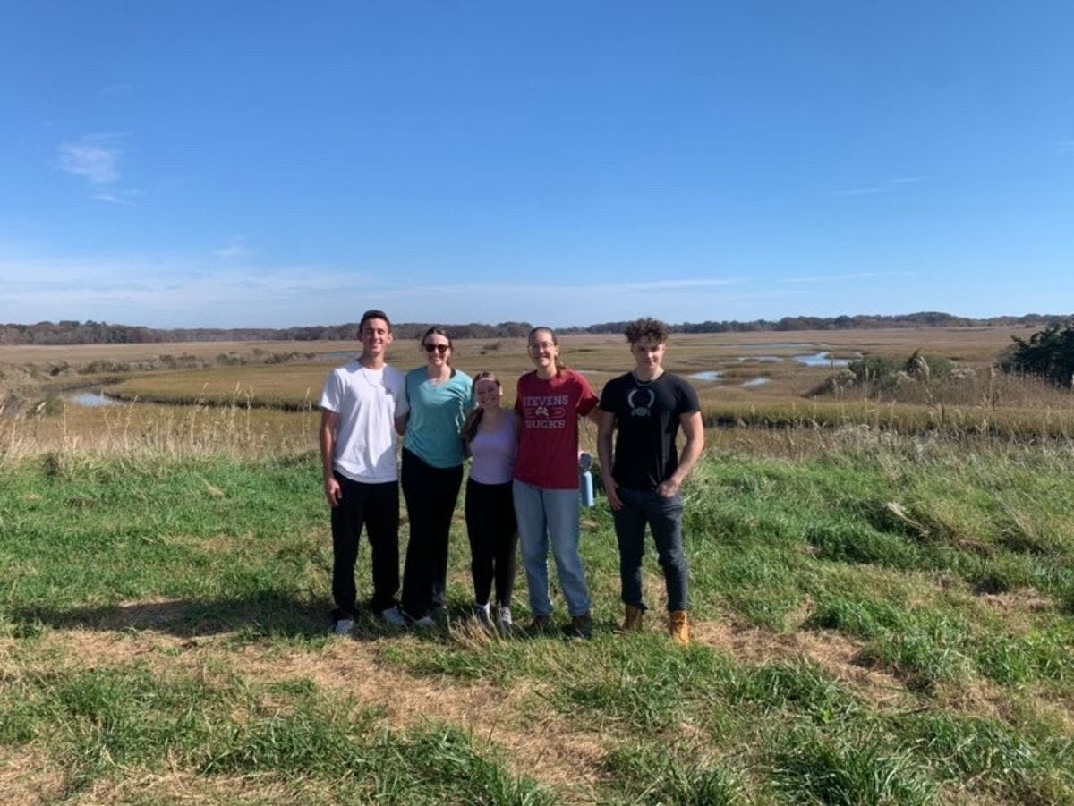A group of five students stand in grass with a meandering creek behind them, at Higbee Beach