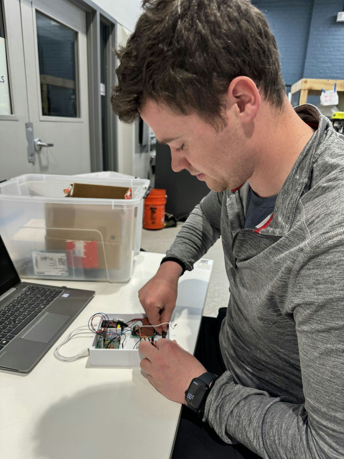 A male student in a gray sweater sits at a desk in a lab. On the desk is an open laptop. In his hands, the student is assembling the MedFlex device.