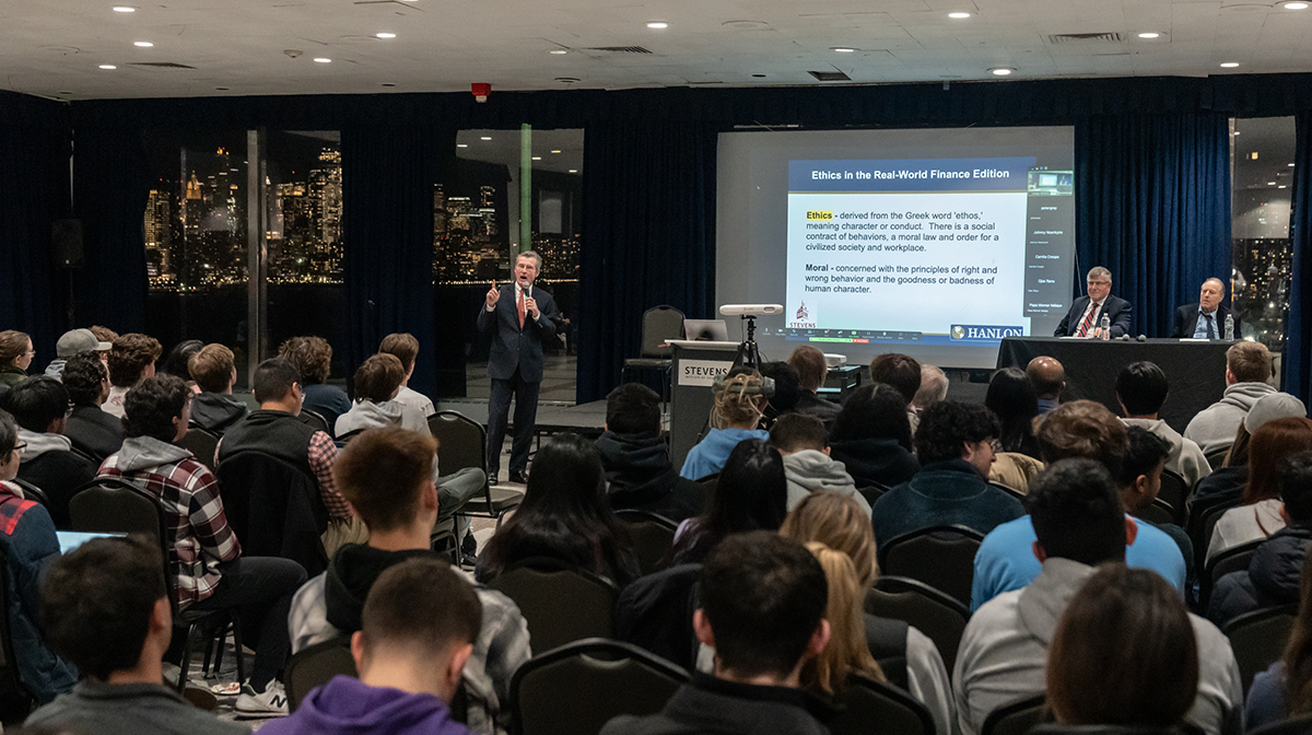 Sean Hanlon speaks to a large crowd of students, faculty and staff. In the background, a partial view of the New York City skyline lit up at night can be seen. 