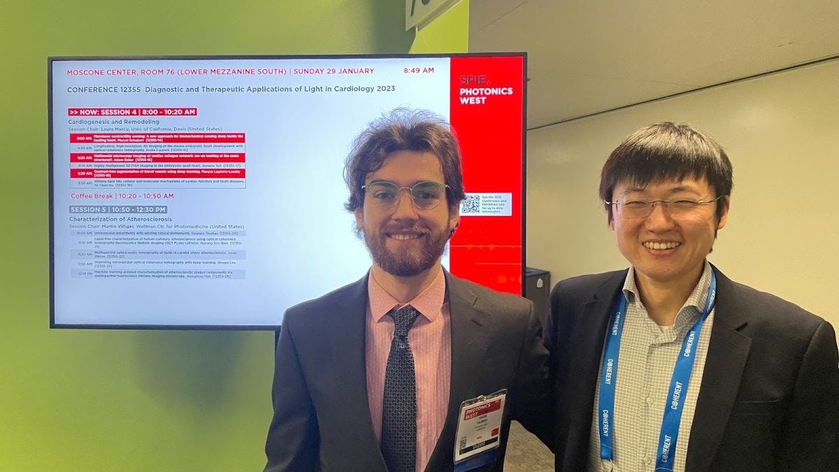 Andre Faubert and Shang Wang standing in front of list of conference proceedings on a television screen
