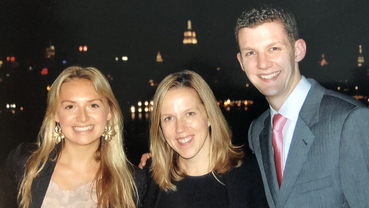 Two women and a man, in formal attire, before the New York City skyline at night.