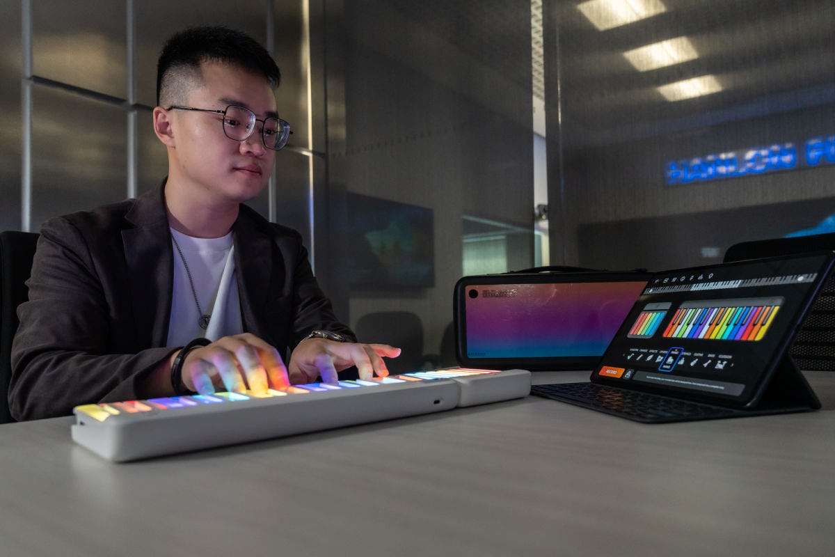 Bohan Zhang sits at a table and plays his PopuPiano keyboard that lights up in front. An iPad sits in front showing the notes he is playing.