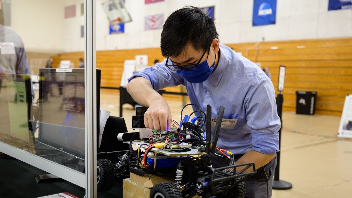 A student wearing a black mask and blue shirt. He is leaning over a robotic vehicle with wheels and adjusting some components on the top of it.