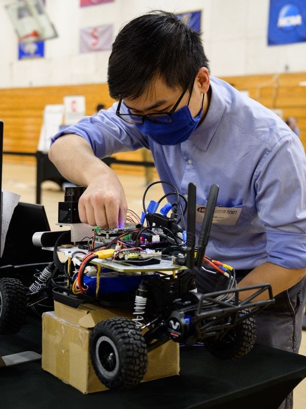 A student wearing a black mask and blue shirt. He is leaning over a robotic vehicle with wheels and adjusting some components on the top of it.