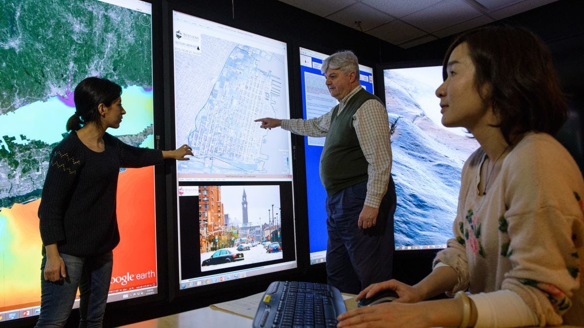 A student in the immersion lab gestures with her hands toward a map on the screen as she's speaking to a professor who is also pointing to the map while another female student looks on
