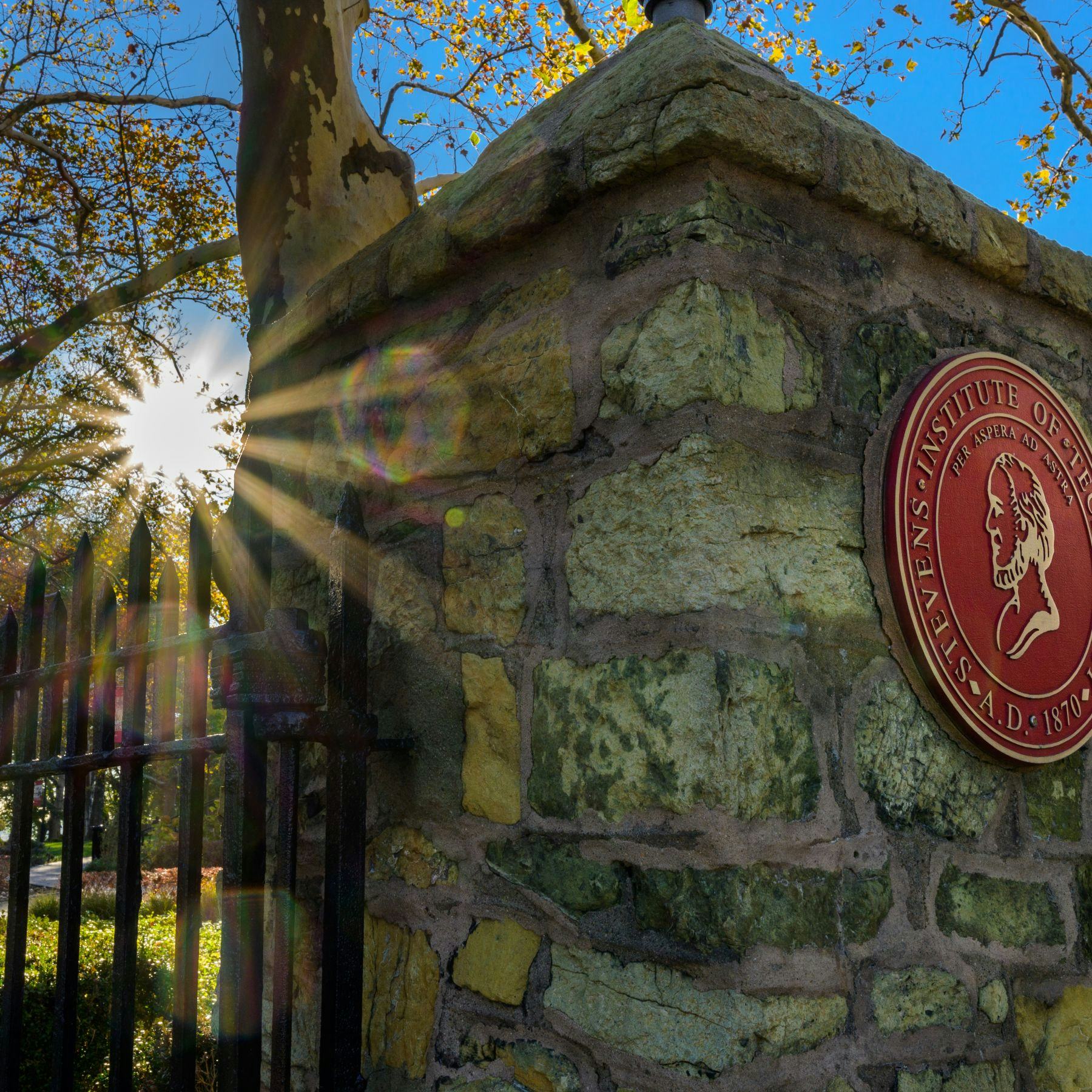 Gateway entrance to Stevens campus with presidents seal and sun shining through trees.