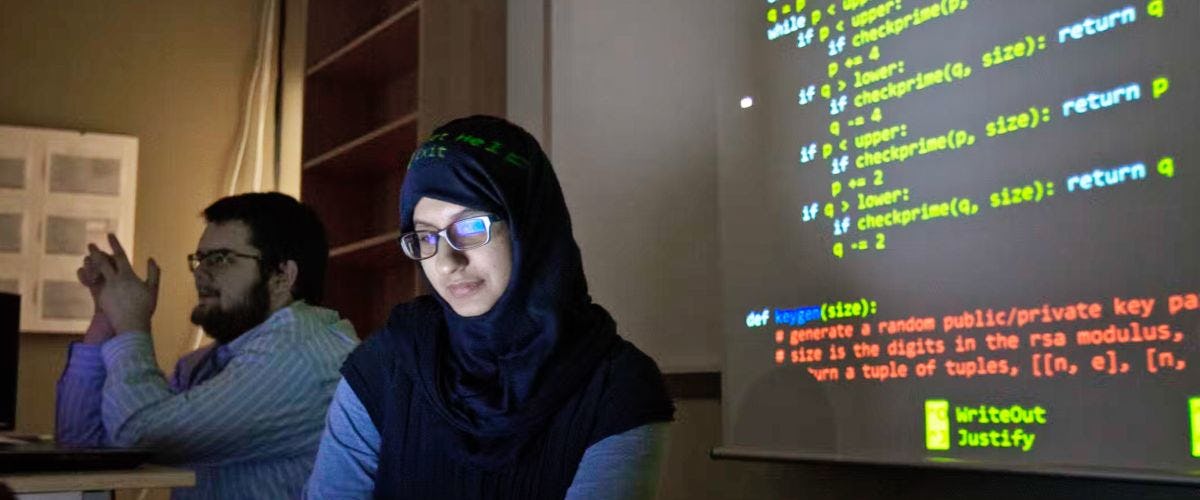 A photo of a female student wearing a hijab working in a dimly lit computer lab with code on the screen in the background