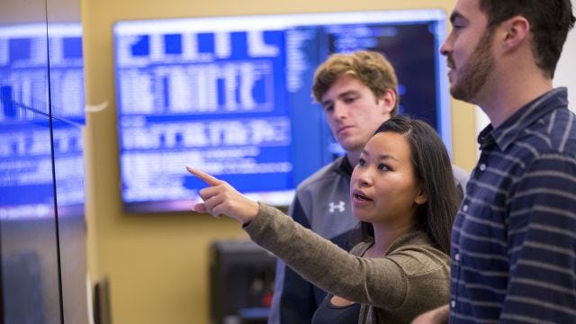 Student points at wall monitor while two other students look on.