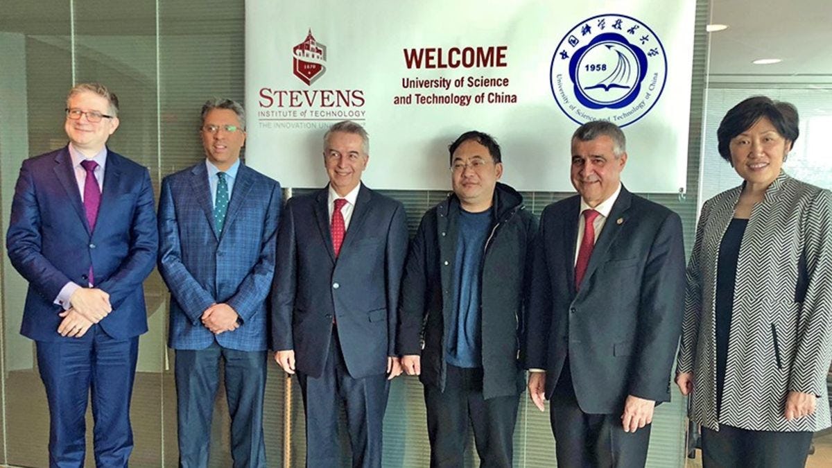 Six people in business attire stand before a banner celebrating the collaboration between Stevens and USTC.