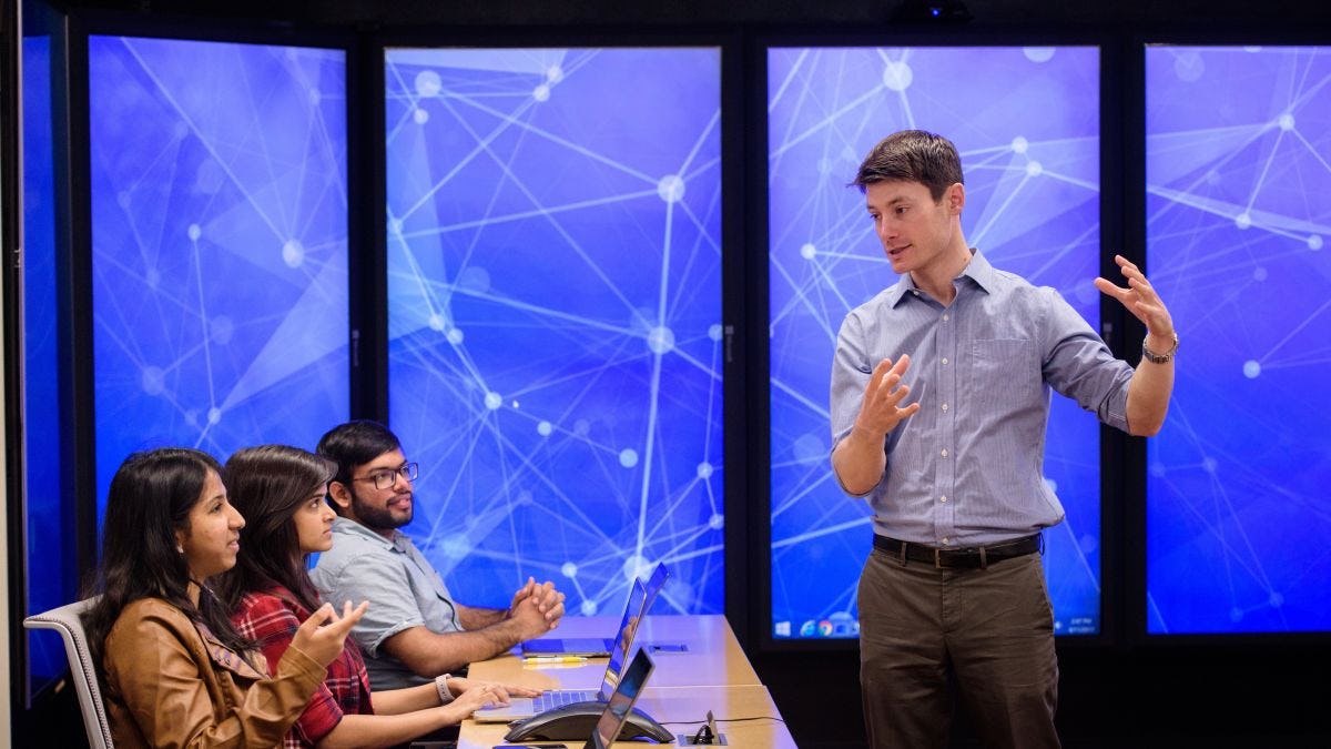 A professor speaks to students in the immersion lab with images of lines and nodes on the screen behind him.