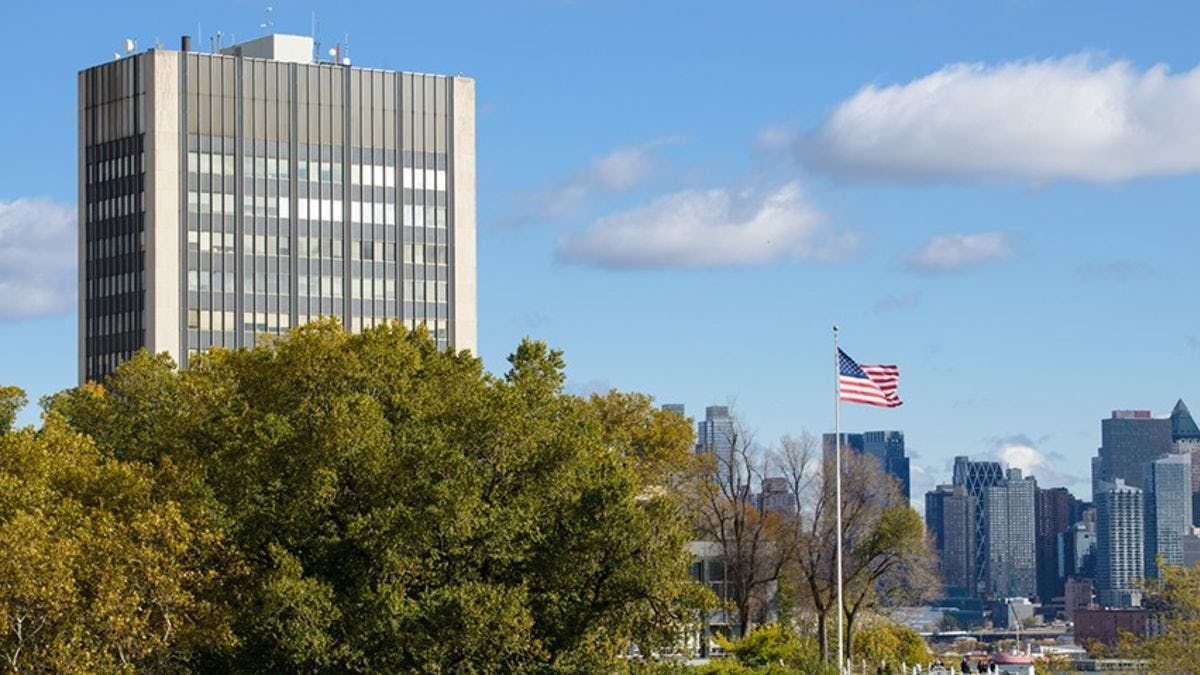 The Howe Center of the Stevens campus at the foreground, with an American flag visible, before the midtown Manhattan skyline. 
