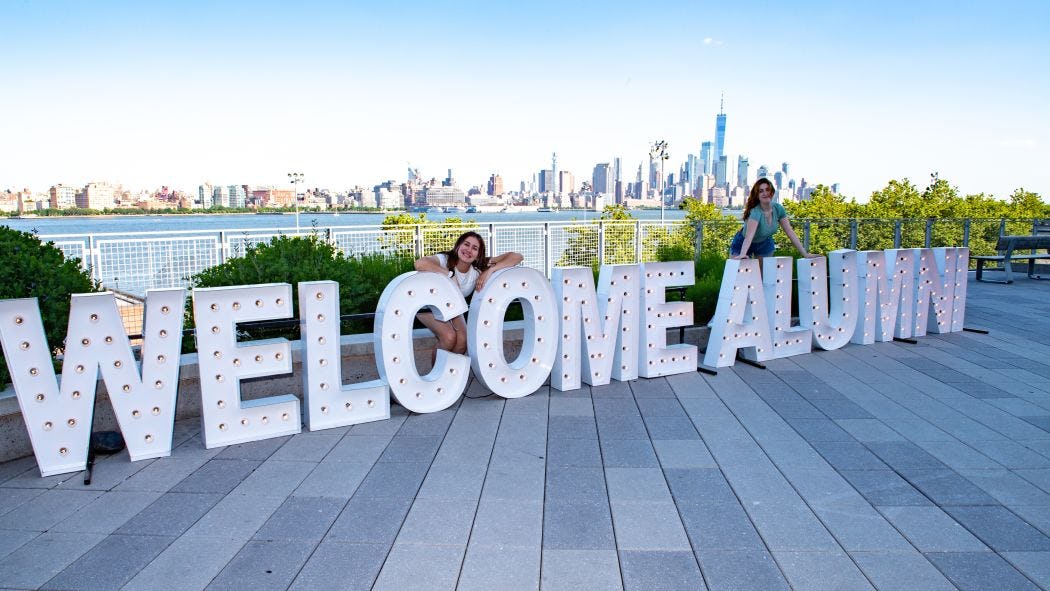 Large letters spelling "Welcome Alumni" on Babbio Patio with two alumnae and NYC skyline in the background