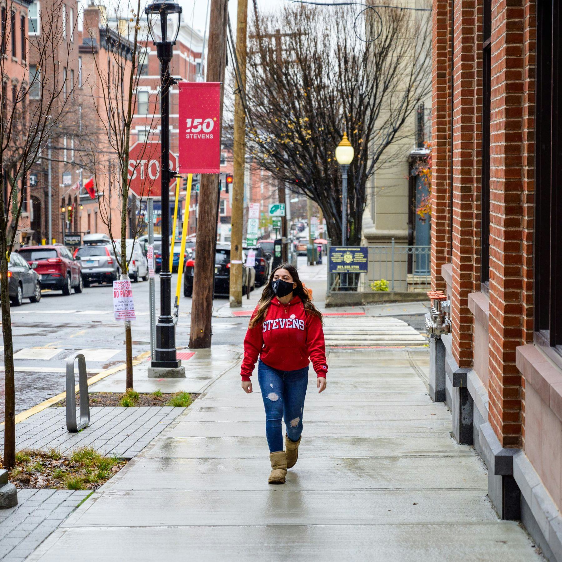 Female Stevens student in red hoodie walking on a Hoboken sidewalk after a rainshower.