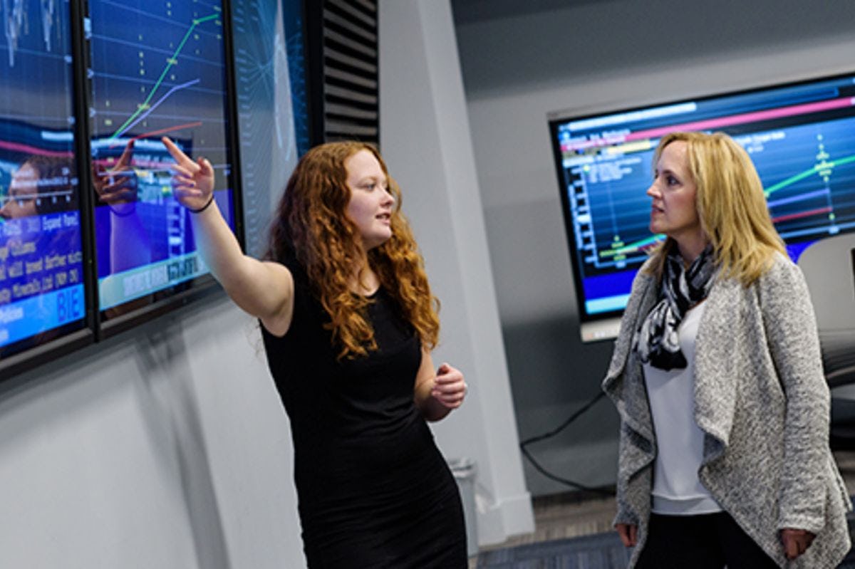 Victoria Piskarev, in a black dress, points to Bloomberg data on a large screen as she presents to Michelle Crilly, in a white sweater.