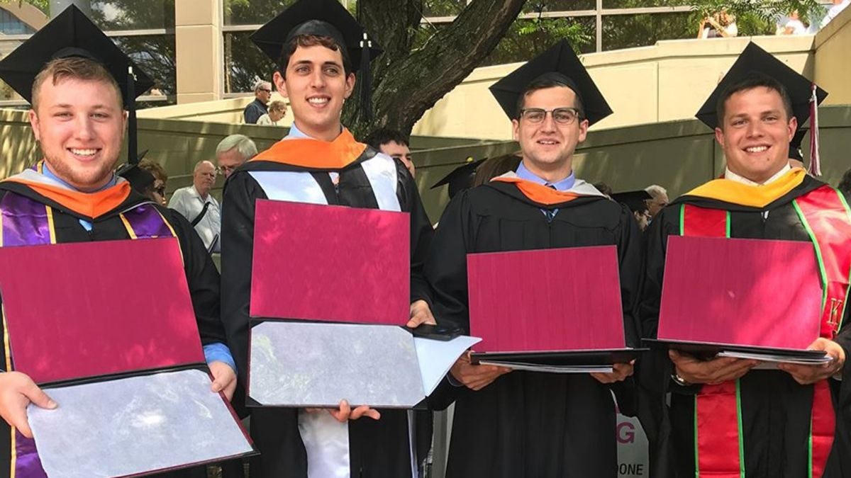 Four men in graduation regalia holding their diplomas as they pose for at commencement. 