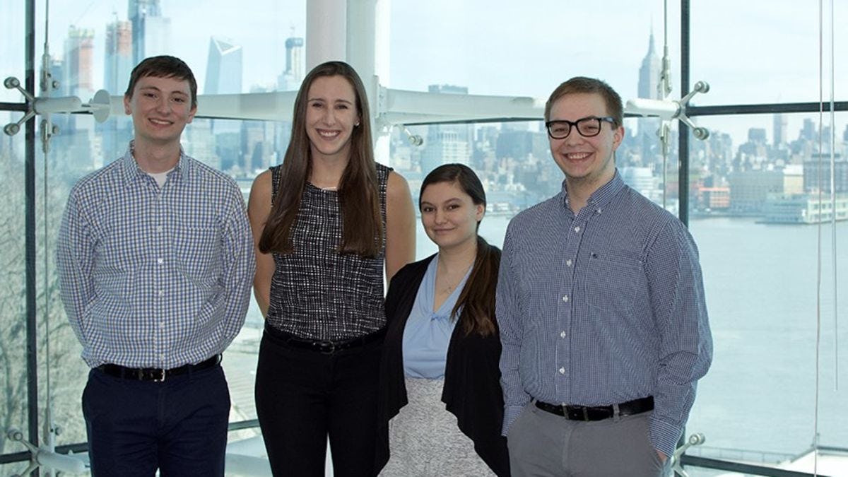 Four students, two men and two women, in a conference room with thw New York City skyline behind them.