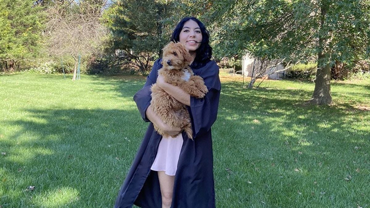 A females student poses in her backyard in graduation regalia while holding her dog.